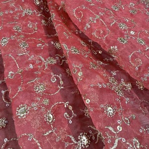 Peut inclure: Gros plan sur un tissu transparent de couleur corail avec une broderie argent&eacute;e d&eacute;taill&eacute;e. La broderie pr&eacute;sente des motifs floraux et tourbillonnants, avec de petits sequins pour plus d'&eacute;clat. Le tissu est drap&eacute;, montrant des plis et de la texture.