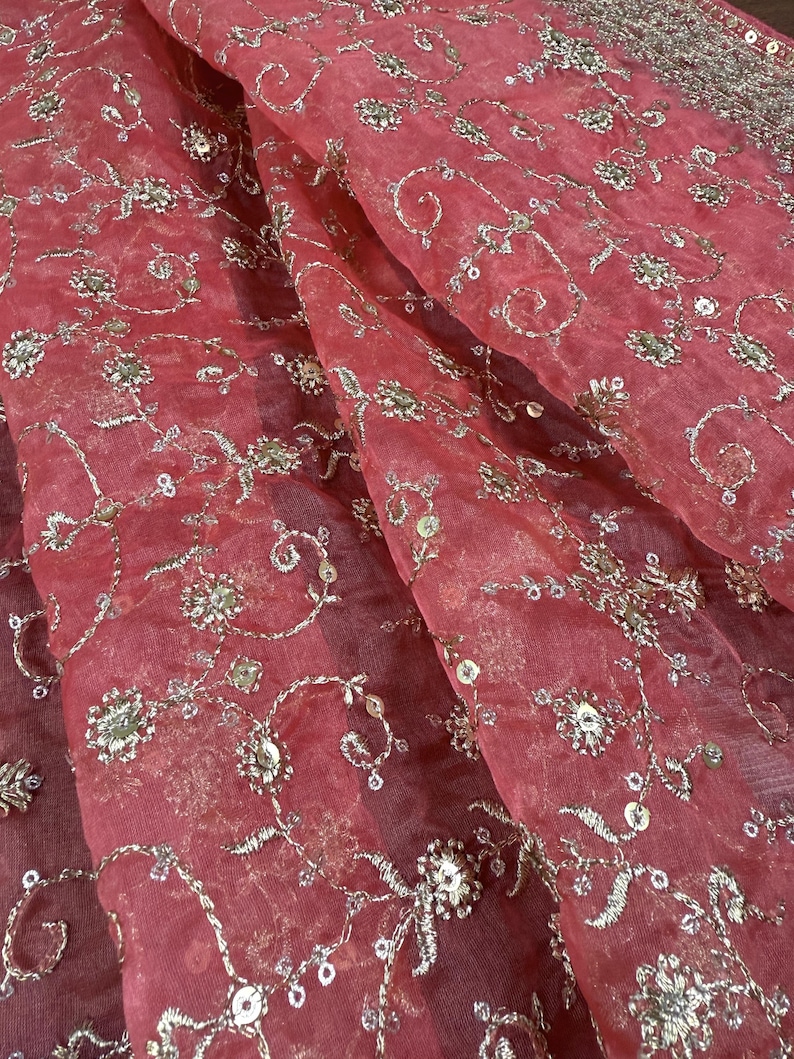 Peut inclure: Gros plan sur un tissu transparent de couleur corail avec une broderie argent&eacute;e d&eacute;taill&eacute;e. La broderie pr&eacute;sente des motifs floraux et tourbillonnants, avec de petits sequins pour plus d'&eacute;clat. Le tissu est drap&eacute;, montrant des plis et de la texture.