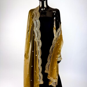 May include: A sheer, golden-yellow shawl with intricate gold embroidery and embellishments. The shawl is draped over a black dress on a mannequin, showcasing the detailed border and delicate fabric. The design features floral patterns.