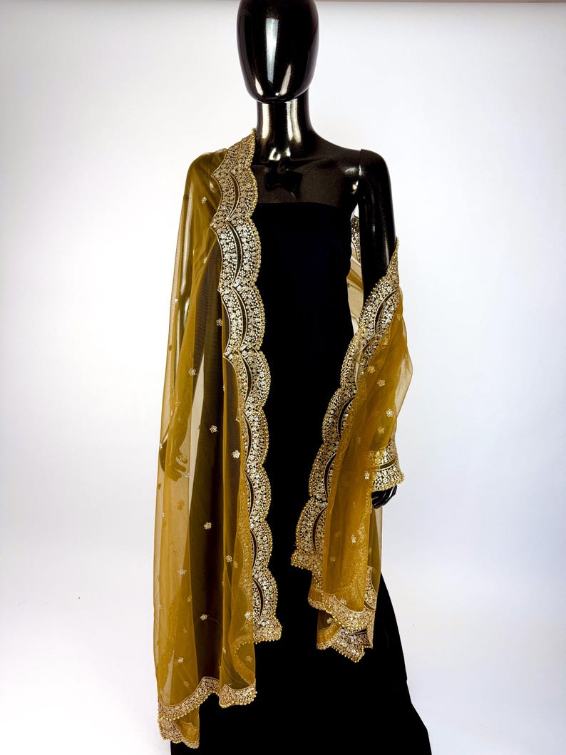 May include: A sheer, golden-yellow shawl with intricate gold embroidery and embellishments. The shawl is draped over a black dress on a mannequin, showcasing the detailed border and delicate fabric. The design features floral patterns.