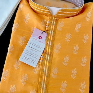 May include: A yellow kurta with a white floral pattern and a white trim around the collar and sleeves. The kurta has a button closure at the neck and a small tag with the text "KAASH COLLECTION" and contact information.