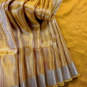 Może przedstawiać: Zbliżenie na drapowaną, mieniącą się tkaninę w odcieniach żółci i złota. Materiał ma pionowe paski i srebrną obwódkę z geometrycznym wzorem. Tkanina jest złożona, tworząc cienie i fakturę.