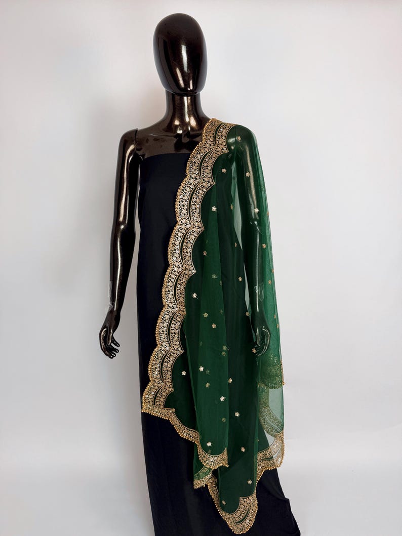 Peut inclure: Une robe noire est drap&eacute;e d'une &eacute;charpe verte transparente. L'&eacute;charpe pr&eacute;sente une bordure dor&eacute;e festonn&eacute;e avec des broderies complexes et de petits ornements blancs &eacute;pars. Le mannequin est sombre et brillant.