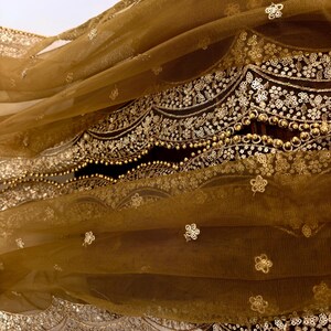 May include: Close-up of a flowing, golden-brown fabric with intricate gold embroidery. The fabric features delicate floral patterns and beaded embellishments along the edges, creating a luxurious and ornate appearance. The sheer material drapes gracefully, showcasing the detailed craftsmanship.