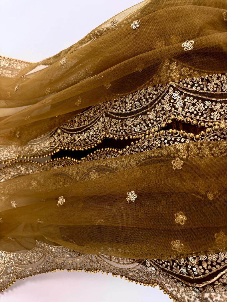 May include: Close-up of a flowing, golden-brown fabric with intricate gold embroidery. The fabric features delicate floral patterns and beaded embellishments along the edges, creating a luxurious and ornate appearance. The sheer material drapes gracefully, showcasing the detailed craftsmanship.
