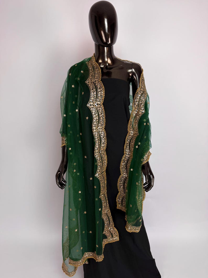 Peut inclure: Un ch&acirc;le vert fonc&eacute; transparent avec une bordure brod&eacute;e dor&eacute;e et des paillettes dor&eacute;es &eacute;parses. Le ch&acirc;le est drap&eacute; sur une robe noire sur un mannequin. La bordure dor&eacute;e a un bord festonn&eacute;.