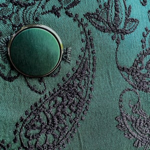 May include: A close-up of a dark green fabric with a black floral pattern. A round, dark green button with a silver rim is in the center of the image.