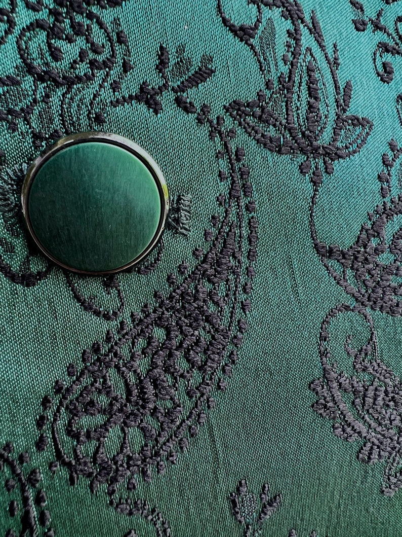 May include: A close-up of a dark green fabric with a black floral pattern. A round, dark green button with a silver rim is in the center of the image.