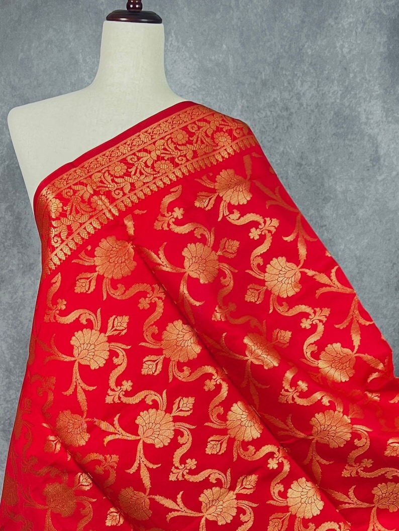 May include: A vibrant red fabric with an intricate gold floral pattern. The fabric is draped over a mannequin, showcasing the detailed design and texture. The border features a decorative gold trim.
