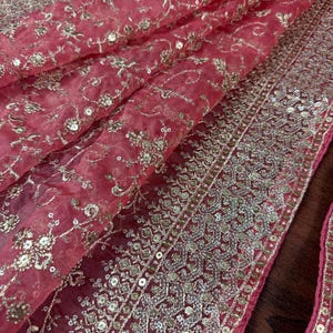 Peut inclure: Gros plan sur un tissu fluide et transparent de couleur rose fonc&eacute;, richement orn&eacute; de broderies et de paillettes dor&eacute;es complexes. Le motif pr&eacute;sente des motifs floraux et g&eacute;om&eacute;triques, cr&eacute;ant un textile luxueux et orn&eacute;.