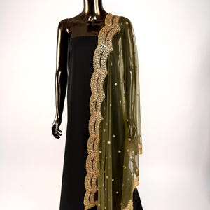 May include: A black dress is draped with a sheer olive green dupatta. The dupatta features a scalloped gold border with intricate embroidery and small gold embellishments. The garment is displayed on a mannequin.