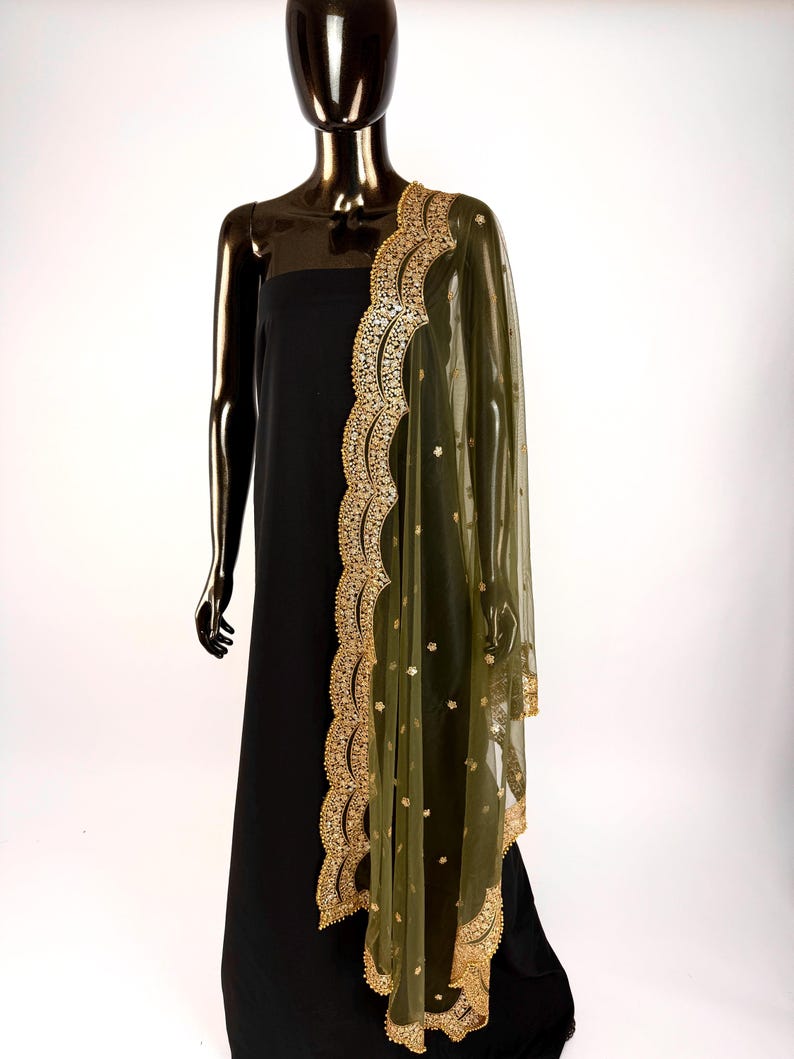 May include: A black dress is draped with a sheer olive green dupatta. The dupatta features a scalloped gold border with intricate embroidery and small gold embellishments. The garment is displayed on a mannequin.