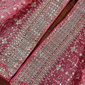 Peut inclure: Gros plan sur un tissu rouge transparent avec des broderies argent&eacute;es et des paillettes complexes. Le motif pr&eacute;sente des motifs floraux et g&eacute;om&eacute;triques sur les bords, cr&eacute;ant une apparence luxueuse et orn&eacute;e. Le tissu est pli&eacute;, r&eacute;v&eacute;lant plusieurs couches.