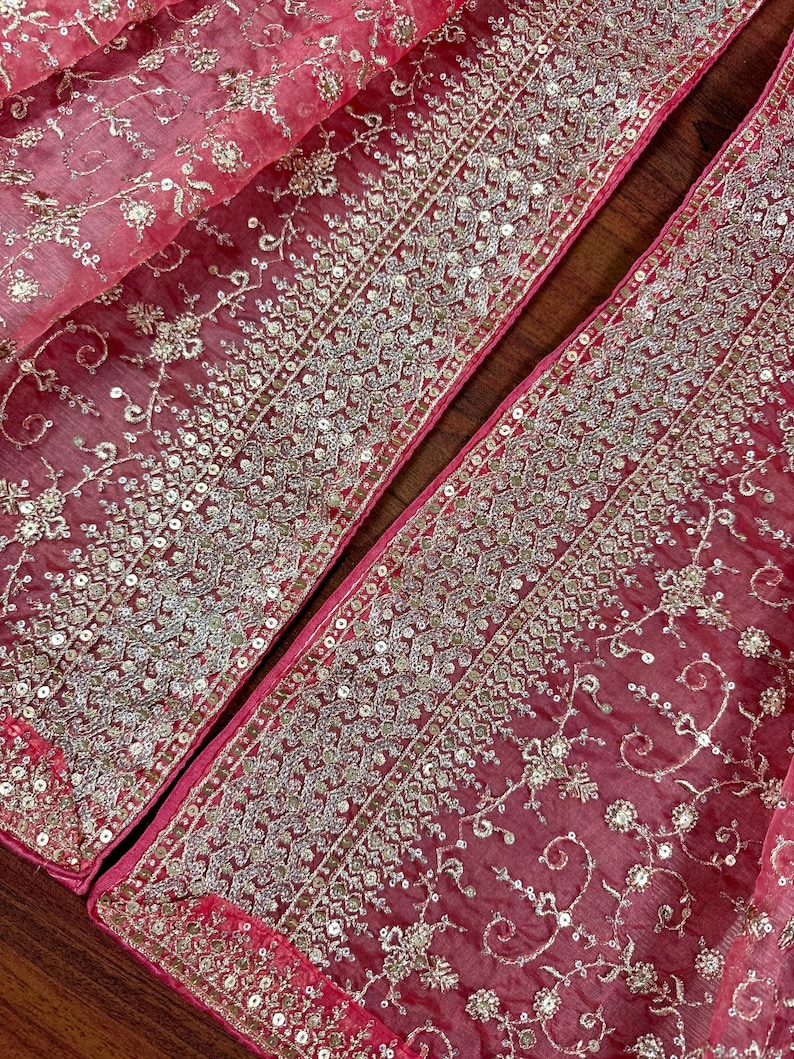 Peut inclure: Gros plan sur un tissu rouge transparent avec des broderies argent&eacute;es et des paillettes complexes. Le motif pr&eacute;sente des motifs floraux et g&eacute;om&eacute;triques sur les bords, cr&eacute;ant une apparence luxueuse et orn&eacute;e. Le tissu est pli&eacute;, r&eacute;v&eacute;lant plusieurs couches.