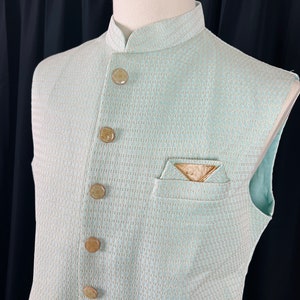 May include: A light blue sleeveless vest with a white and gold patterned design. The vest has a mandarin collar and six gold buttons down the front.