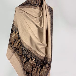 Op de afbeelding: Een beige sjaal met een brede zwarte rand versierd met ingewikkeld goudkleurig borduurwerk. Het patroon toont een processie van figuren en olifanten in traditionele kleding, met gedetailleerde motieven die langs de randen van de stof lopen.
