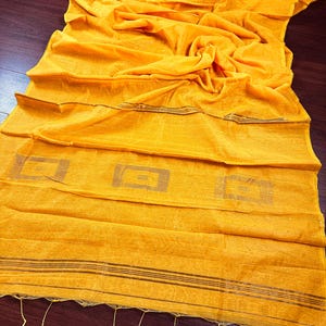 Puede incluir: Un chal o bufanda de color amarillo brillante con un tejido texturizado. La tela está drapeada, mostrando pliegues y capas. El borde inferior tiene borlas decorativas. El diseño incluye rayas horizontales y formas geométricas.