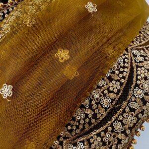 May include: Close-up of a gold and black textile with intricate embroidery and embellishments. A sheer, mustard-colored fabric with floral accents overlays the detailed design. The textile features sequins and beaded trim.