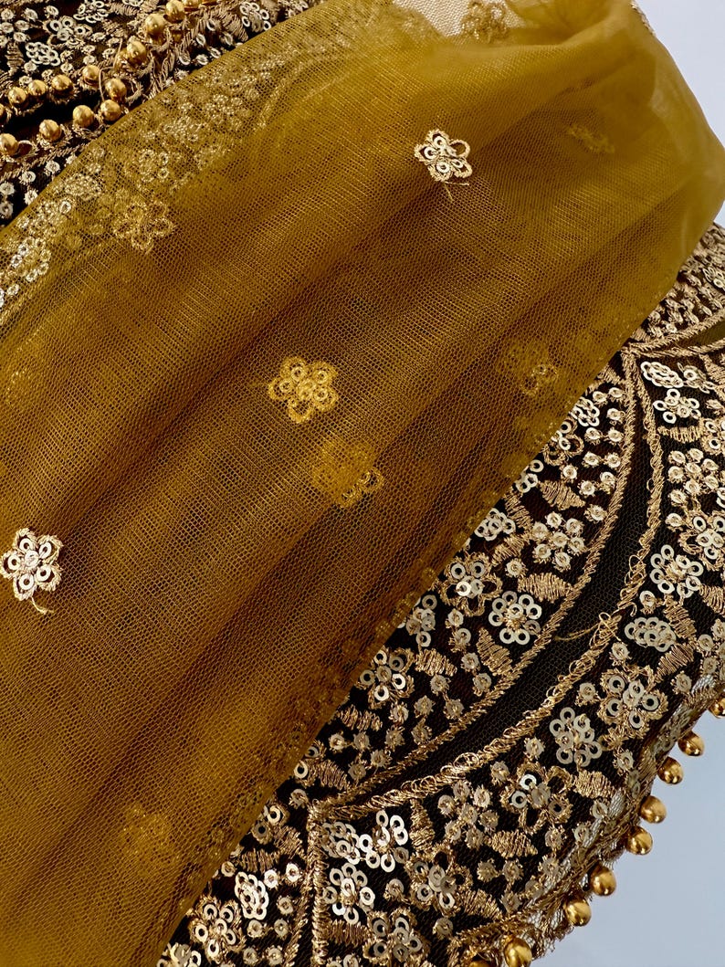 May include: Close-up of a gold and black textile with intricate embroidery and embellishments. A sheer, mustard-colored fabric with floral accents overlays the detailed design. The textile features sequins and beaded trim.