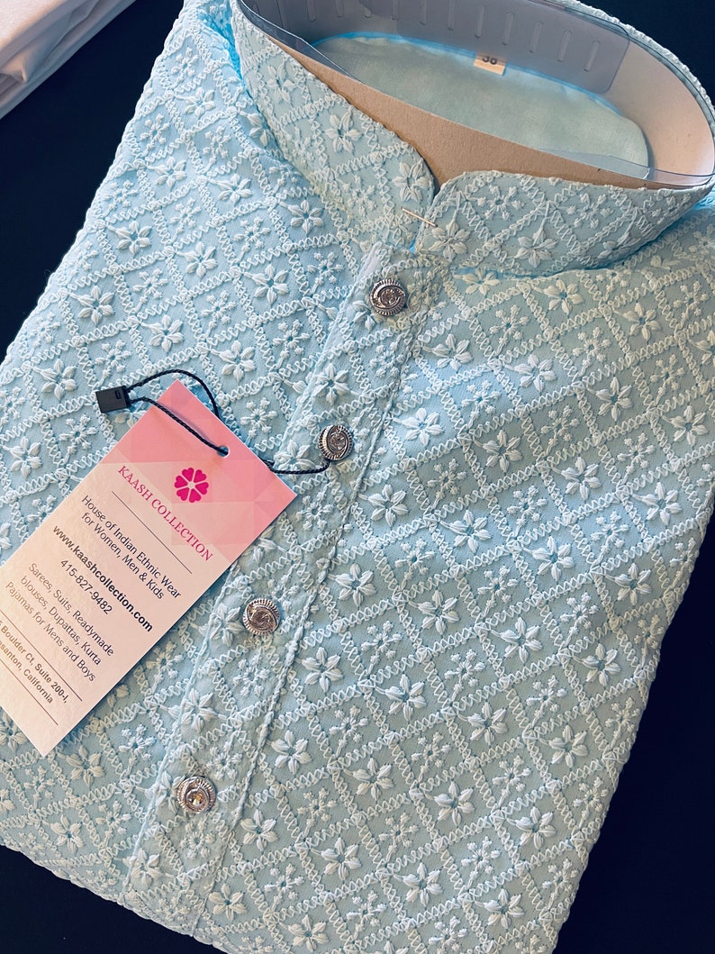 Pu&ograve; includere: Una kurta blu chiaro ricamata con un motivo floreale bianco. La kurta ha un colletto alla coreana e chiusura con bottoni. L'etichetta recita "Kaash Collection, House of Indian Ethnic Wear for Women, Men & Kids, www.kaashcollection.com, 415-827-9482".