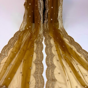 May include: A sheer, golden-brown shawl with intricate gold embroidery along the edges. The scalloped borders are embellished with small, sparkling accents. The delicate fabric drapes gracefully, showcasing the detailed craftsmanship.