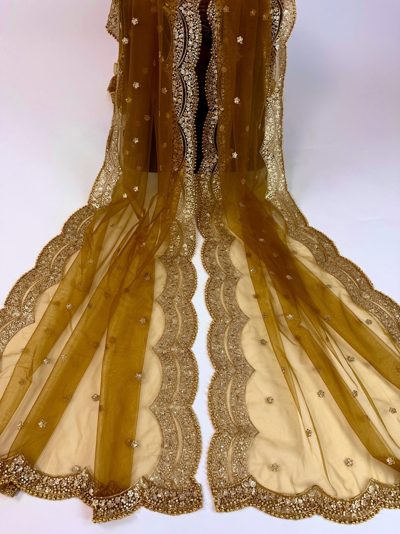 May include: A sheer, golden-brown shawl with intricate gold embroidery along the edges. The scalloped borders are embellished with small, sparkling accents. The delicate fabric drapes gracefully, showcasing the detailed craftsmanship.
