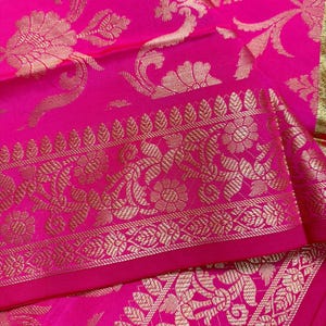 May include: A vibrant hot pink fabric featuring an intricate gold metallic woven pattern. The design includes detailed floral motifs and scrolling vines, with a prominent decorative border showcasing repeating leaf and flower arrangements.