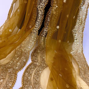 May include: Mustard-colored sheer fabric with intricate gold embellishments. The fabric features scalloped edges adorned with sequins and small gold beads. Delicate floral patterns are scattered across the sheer material, creating a luxurious and ornate design.