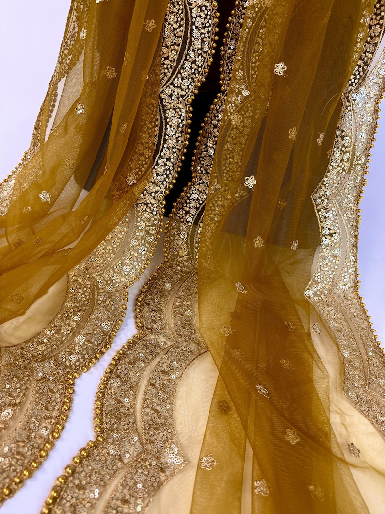 May include: Mustard-colored sheer fabric with intricate gold embellishments. The fabric features scalloped edges adorned with sequins and small gold beads. Delicate floral patterns are scattered across the sheer material, creating a luxurious and ornate design.