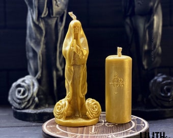 May include: Two gold-coloured candles. One is a figure of a robed person with hands clasped in prayer, and the other is a cylindrical pillar candle with an embossed design. Both candles are on a wooden platform with etched symbols. The background features dark statues.