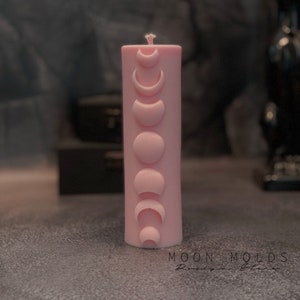 May include: A pink pillar candle with a design of eight moon phases. The candle is decorated with a white wax design of the moon phases.