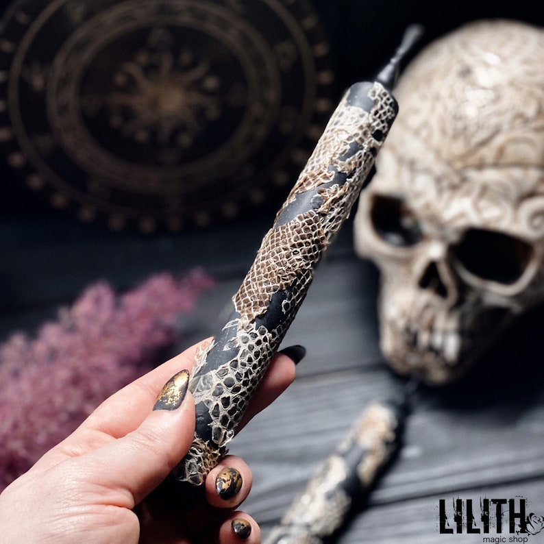May include: A black and brown candle with a snakeskin pattern. The candle is held in a hand with gold nail polish.