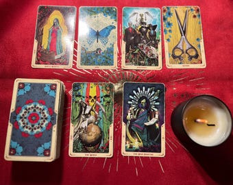 May include: Tarot card spread with ten cards, including Death, The World, The High Priestess, and Justice. The cards are laid out in a cross pattern on a red fabric surface. A lit candle is in the lower right corner of the image.