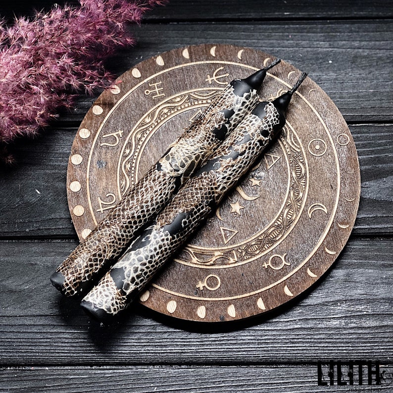 May include: Two black candles with a snake skin pattern on a wooden circular base with astrological symbols engraved on it.