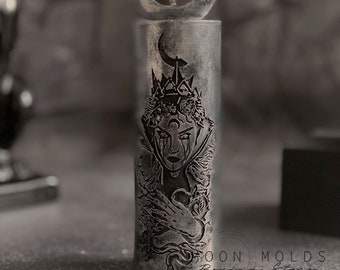 May include: A silver candle with a black and white design of a woman with a crown and a snake. The candle has a crescent moon on top and the text "Moon Molds Design Studio" at the bottom.