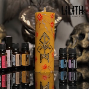 Könnte beinhalten: Eine hohe, gelbe Säulenkerze mit einem schwarzen und goldenen Symbol und roten Blumenakzenten. Kleine Flaschen mit ätherischen Ölen umgeben die Kerze. Der Text "LILITH magic shop" ist sichtbar.