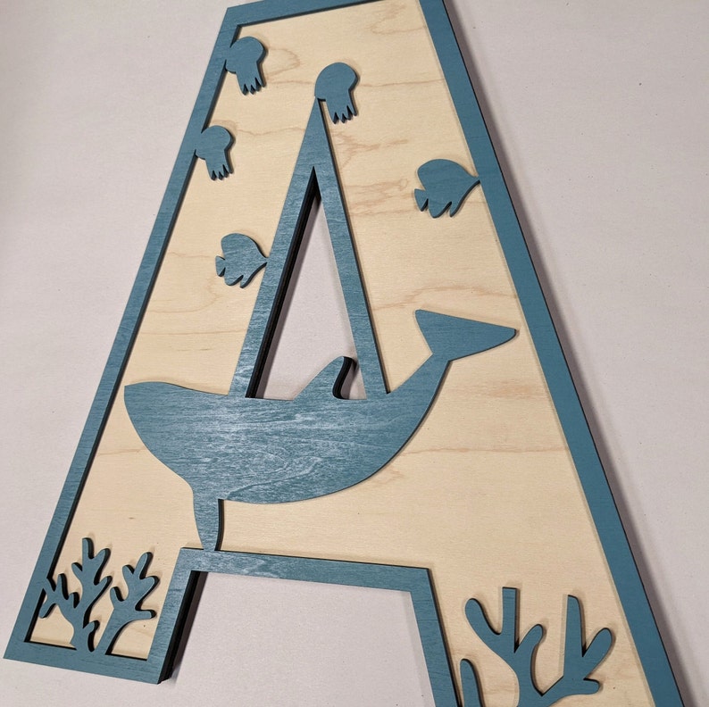 Op de afbeelding: Een houten letter A geverfd in een lichtblauwe kleur. De letter is versierd met een walvis, een zeilboot en koraal. De walvis springt uit het water en de zeilboot vaart over het water. Het koraal bevindt zich onderaan de letter.