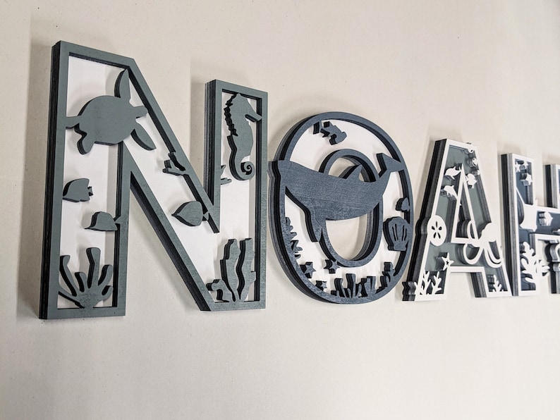 Op de afbeelding: Grijze houten letters die "NOAH" spellen met een nautisch thema. Elke letter is versierd met zeedieren, zeewier en andere ocean-thema ontwerpen.