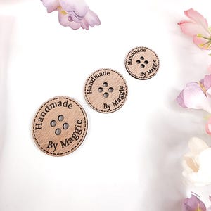 May include: Three wooden buttons of different sizes, each with four holes and the text "Handmade By Maggie" etched around the edge. The buttons are light brown with a dark brown stitched border. The background includes flowers and fabric.