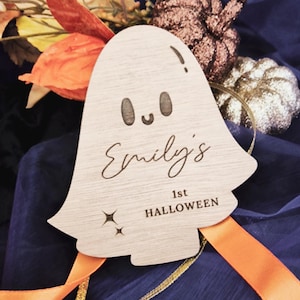 May include: A wooden ghost-shaped decoration with the text "Emily's 1st Halloween." The ghost has a simple face and star accents. The decoration is set against a backdrop of orange and gold ribbons and fall foliage.