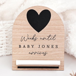 May include: Wooden baby countdown sign with a black heart at the top. The sign reads "Weeks until BABY JONES arrives" in a script font. A white chalk stick and a black stripe are at the bottom. The sign is made of light-coloured wood.