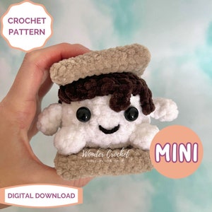 May include: A crocheted amigurumi s'more with a brown chocolate bar, white marshmallow, and two graham cracker pieces. The s'more has a smiling face and black eyes. The s'more is labeled "MINI".