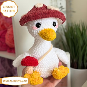 Peut inclure: Un canard en crochet blanc fait &agrave; la main portant un chapeau champignon bordeaux et tenant un champignon rouge. Le canard a un bec et des pieds jaunes, avec des joues roses. Le texte "CROCHET PATTERN" et "DIGITAL DOWNLOAD" est inclus.