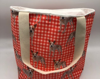 May include: A red and white gingham tote bag with a print of Australian Cattle Dogs. The bag has two cream-colored canvas straps.