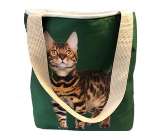 May include: Green canvas tote bag with a brown and black tabby cat printed on the front. The bag has cream colored straps.