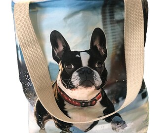 May include: A canvas tote bag featuring a Boston Terrier dog in a winter scene. The dog is black and white with a red collar. The bag has beige straps and a blue and white background.