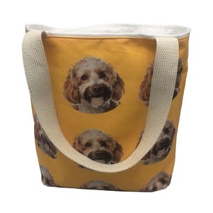 Könnte beinhalten: Eine gelbe Canvas-Tragetasche mit weißen Henkeln. Die Tasche hat ein sich wiederholendes Muster eines Hundefaces.