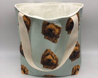 May include: A light blue tote bag featuring a repeating pattern of a brown dog's face. The bag has cream-colored handles and a cream-colored interior lining. The bag is made of a sturdy fabric.
