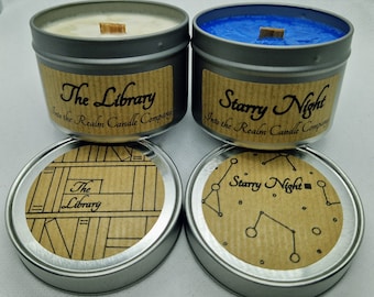May include: Two candles in silver tins with brown paper labels. The candle on the left is labeled "The Library" and the candle on the right is labeled "Starry Night". Both candles are from "Into the Realm Candle Company".