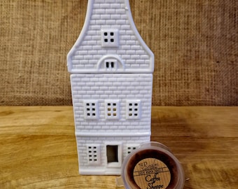May include: A white ceramic house-shaped wax warmer with a brick pattern, featuring multiple windows and a door. A container of "Coffee Shoppe" soy wax melt sits in front of the warmer. The warmer is on a wooden surface.